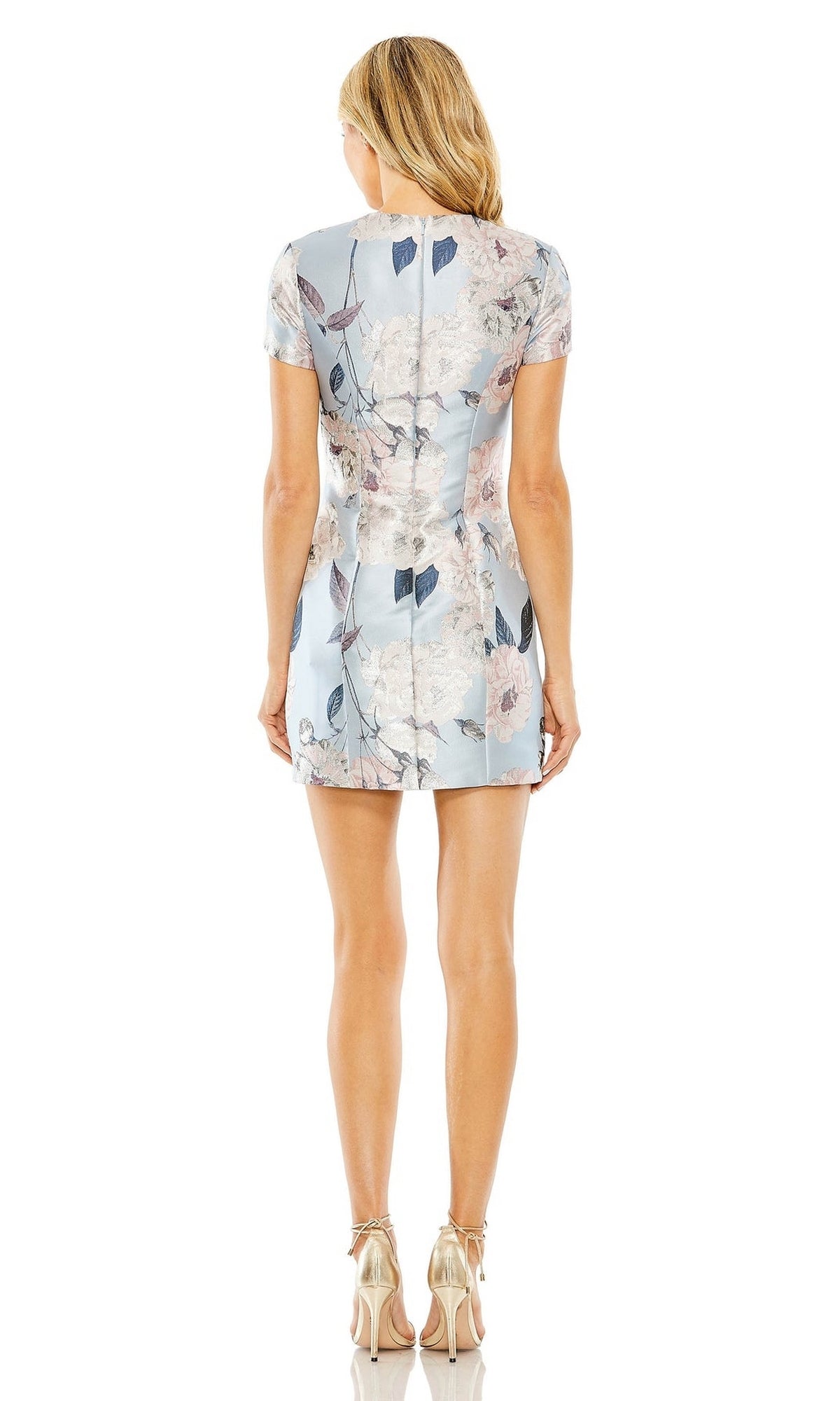 A woman stands facing away, wearing the Mac Duggal Floral-Print Wedding-Guest Dress 11614 in light blue with short sleeves, paired with metallic high-heeled shoes.